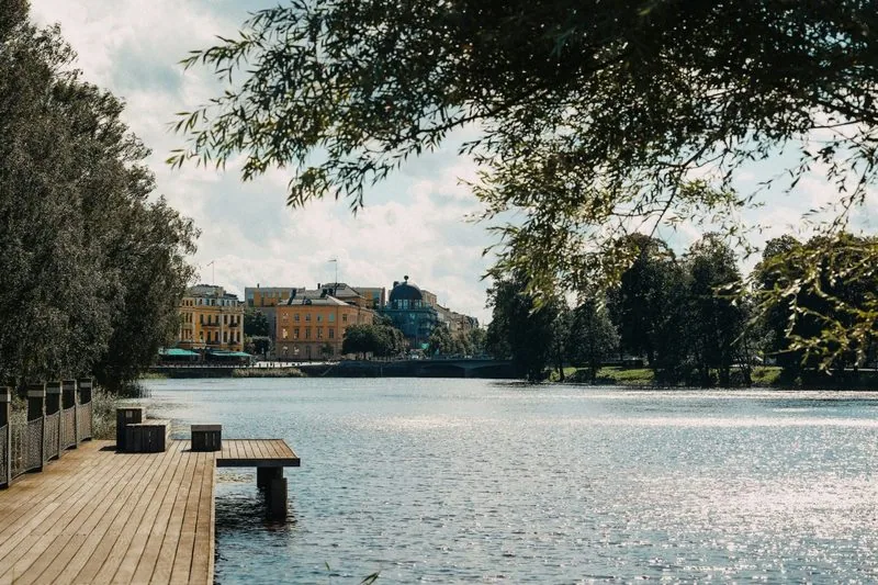 Holzsteg am Flussufer, Weidenzweige im Vordergrund, dahinter eine Häuserzeile mit Kuppelbau