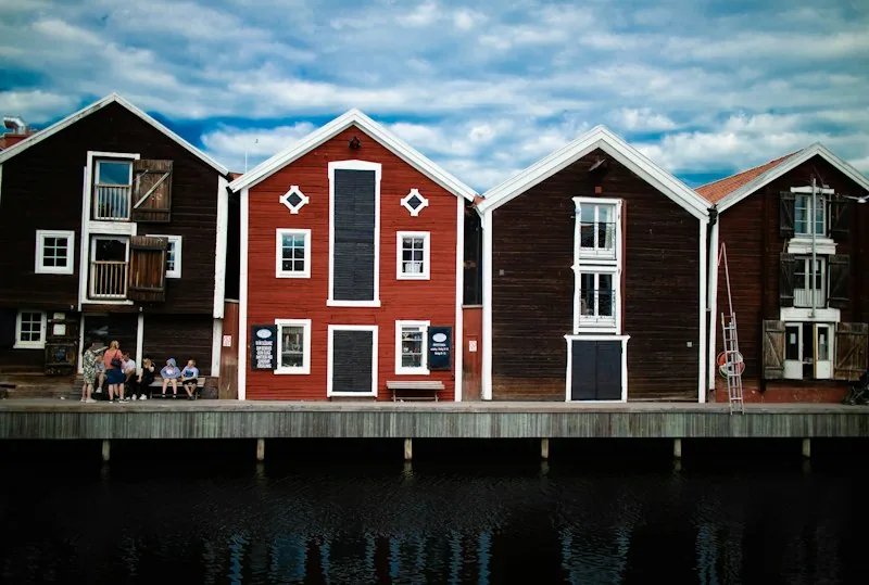 Rote und braune Holzspeicher mit weißen Kanten am Wasser in Hudiksvall, darüber blauer Himmel mit Wolken