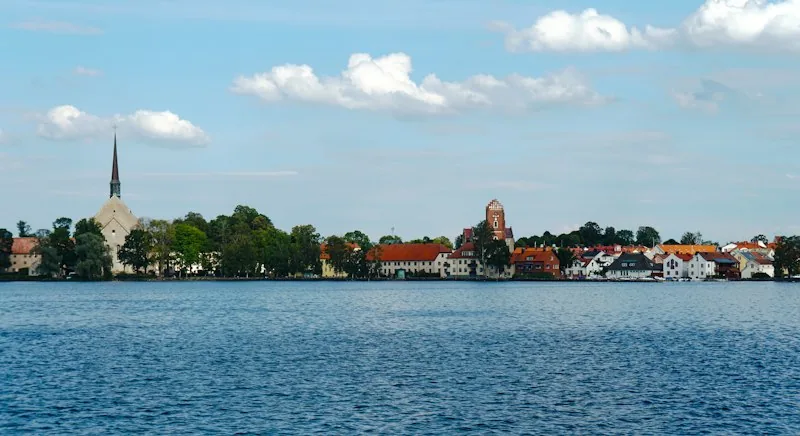 Blick über den Vättern auf Vadstena, rote Dächer und ein Kirchturm hinter den Bäumen am Wasser