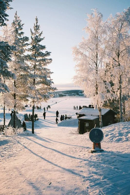 Winterlicher See mit reifbedeckten Birken und Kiefern, Menschen klein auf dem Eis im rosa Winterlicht, eine Holzhütte im Schnee